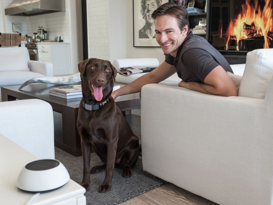 A man with his dog in a living room with a lit fireplace, leaning toward the Josh Micro device.